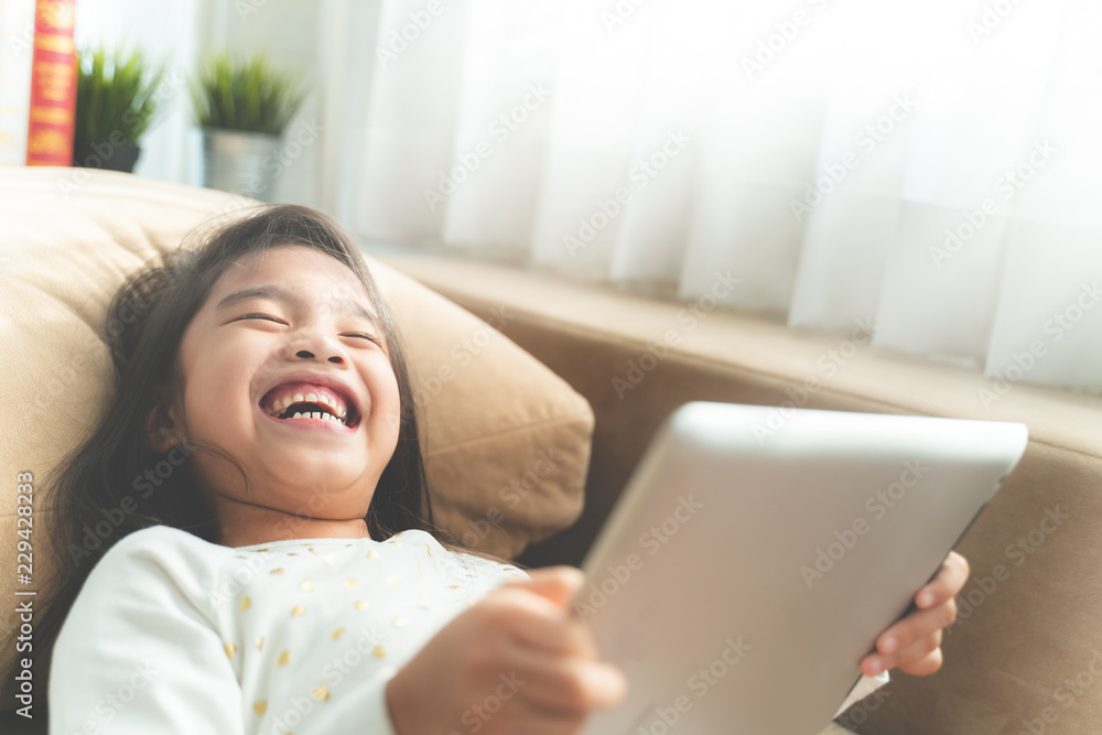 Asian Cute child playing games with a tablet and smiling while sitting on sofa at home Asian Cute child playing games with a tablet and smiling while sitting on sofa at home