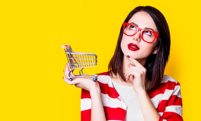 Portrait of a young woman in glasses with shopping cart on yellow background