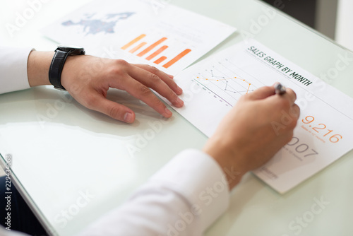 Close-up of male hands writing on paper with graph sale. Caucasian businessman sitting at table and working with marketing papers. Marketing concept