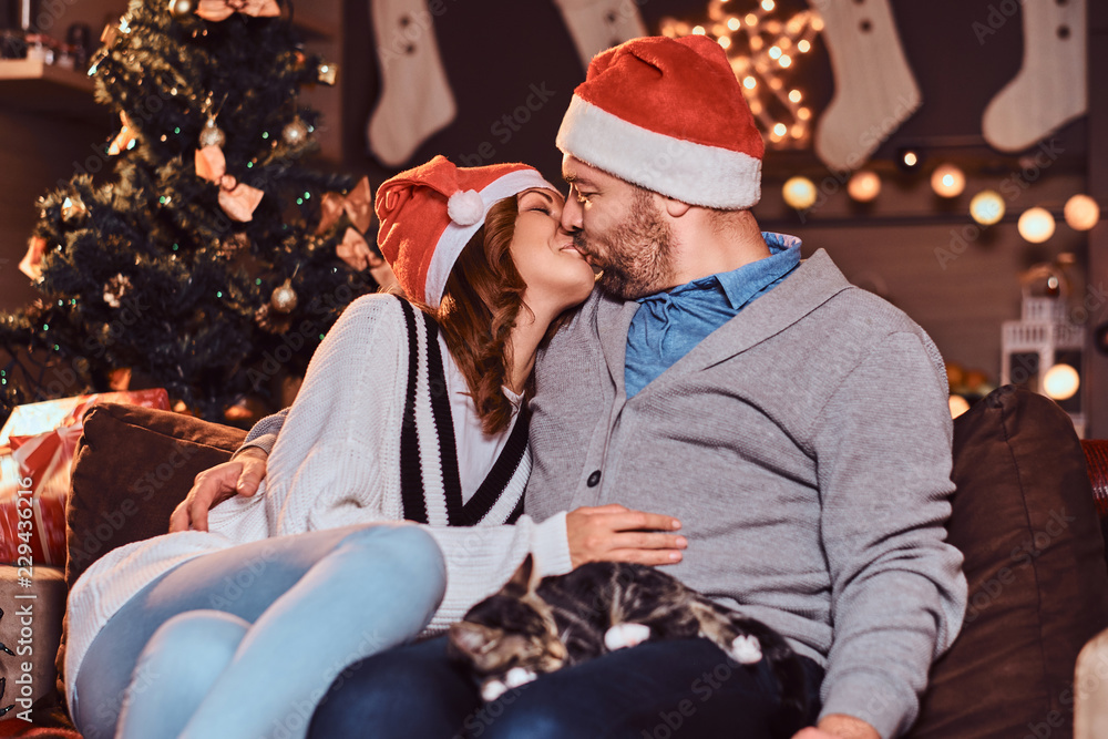 Happy couple celebrating Christmas Eve at home kiss while sitting on sofa. Christmas holiday.