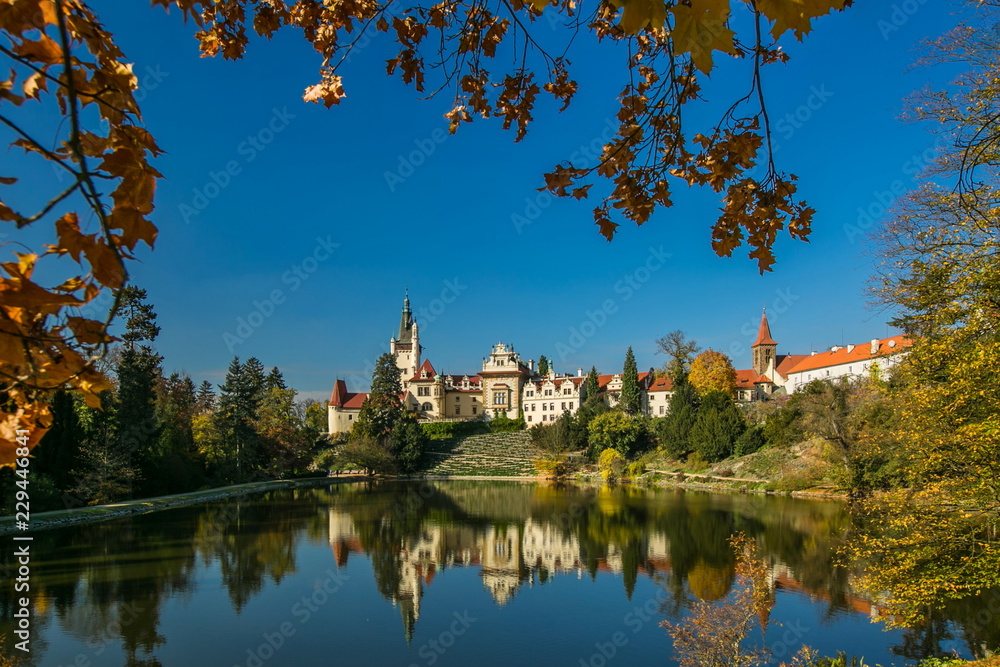 Beautiful fall scenery of famous Pruhonice castle, Czech Republic, Europe, standing on hill, sunny day, blue sky, reflection in lake, yellow maple leaves in foreground, colorful trees