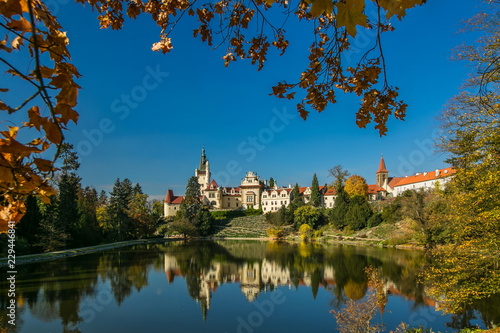 Beautiful fall scenery of famous Pruhonice castle, Czech Republic, Europe, standing on hill, sunny day, blue sky, reflection in lake, yellow maple leaves in foreground, colorful trees