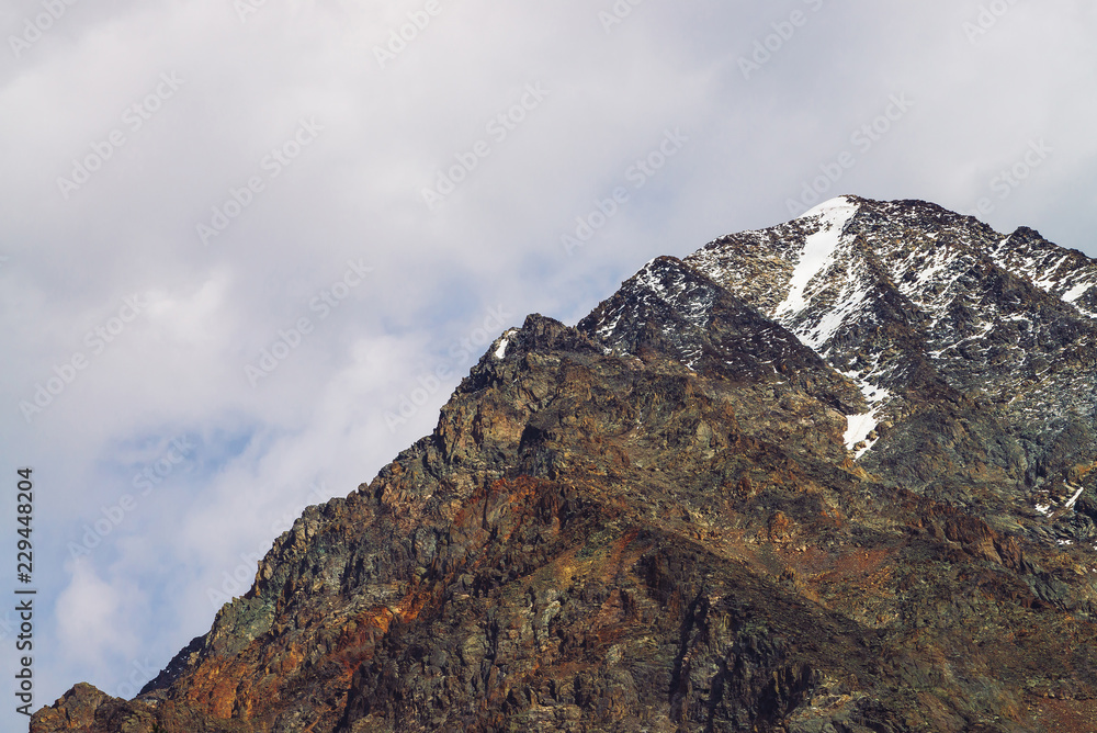 Snowy mountain top in cloudy sky. Rocky ridge under clouds. Overcast weather in highlands. Atmospheric minimalist landscape of majestic nature.