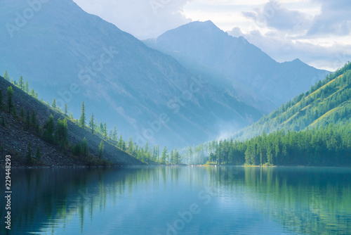 Ghostly mountain lake in highlands at early morning. Beautiful misty mountains reflected in calm clear water surface. Smoke of campfires. Amazing atmospheric foggy landscape of majestic nature.