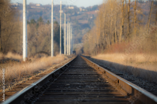 Low level view of train tracks in rural area with foreground in focus, background out focus, power poles and pale blue overcast sky  © Jacquie Klose