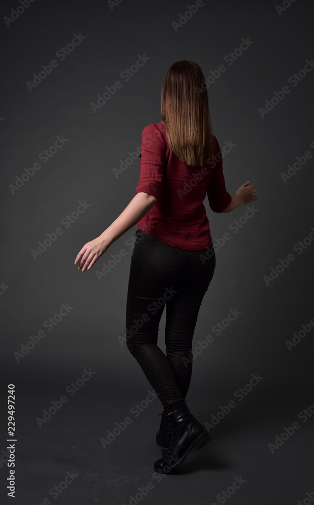 full length portrait of brunette girl wearing red shirt and leather pants. standing pose, facing away from the camera, on grey studio background. full length portrait of brunette girl wearing red shirt and leather pants. standing pose, facing away from the camera, on grey studio background.