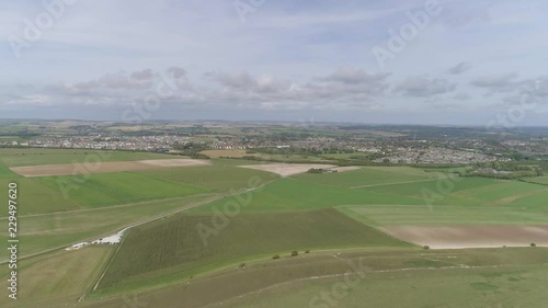 Aerial tracking north from Maiden Castle, the towns of Dorchester and Poundbury is visible in the background. Lots of fields surround the scene. photo