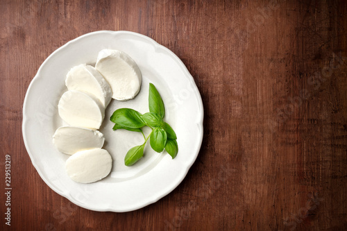 An overhead photo of a white plate with fresh basil leaves and Mozzarella cheese, shot from above on a dark rustic wooden background with a place for text
