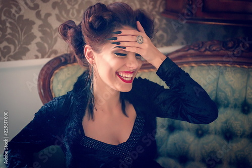 Canvas Print Sexy Caucasian model looking like a vampire laughing on a couch, having sculpting make-up and cocktail dress with big cleveage