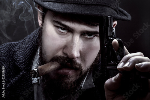 bearded gangster in a hat with a gun in his hand and a cigarette in his mouth on black background