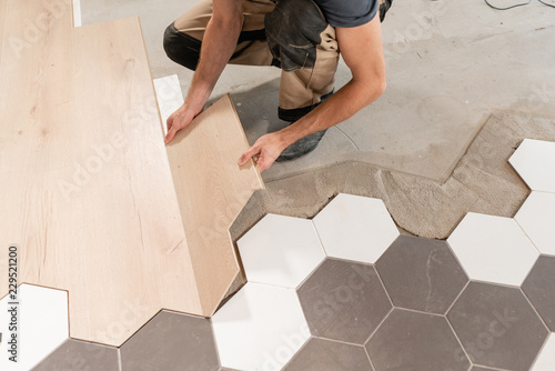 Male worker installing new wooden laminate flooring. The combination of wood panels of laminate and ceramic tiles in the form of honeycomb. Kitchen renovation.