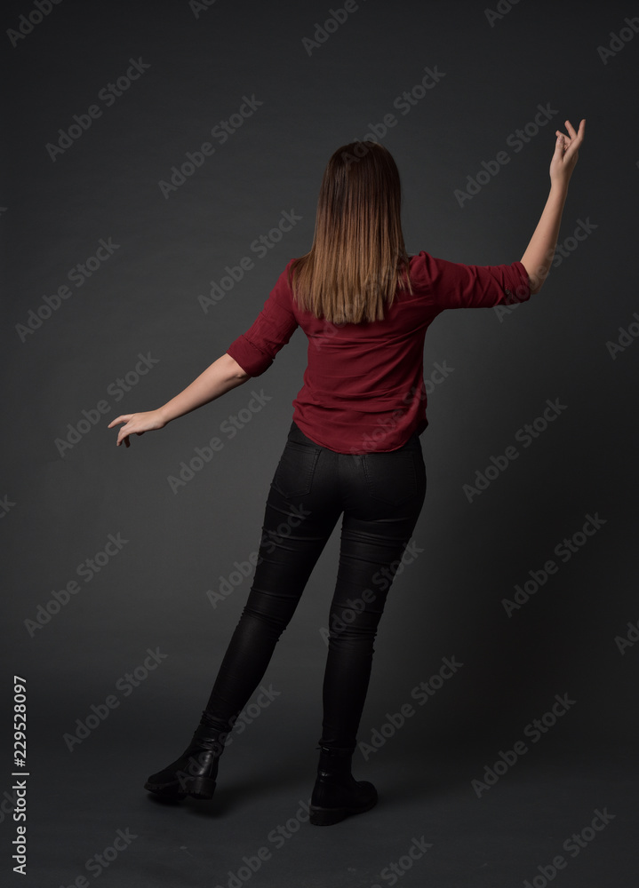 full length portrait of brunette girl wearing  red shirt and leather pants. standing pose, facing away from the camera, on grey studio background.