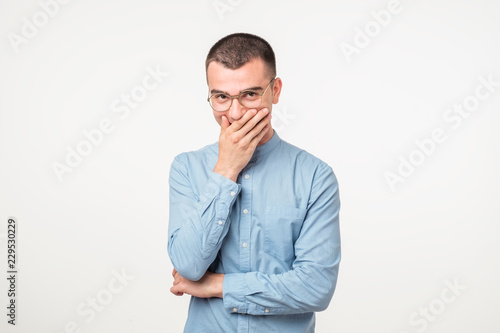 Portrait of young man laughing and covering his mouth with hand over white background.