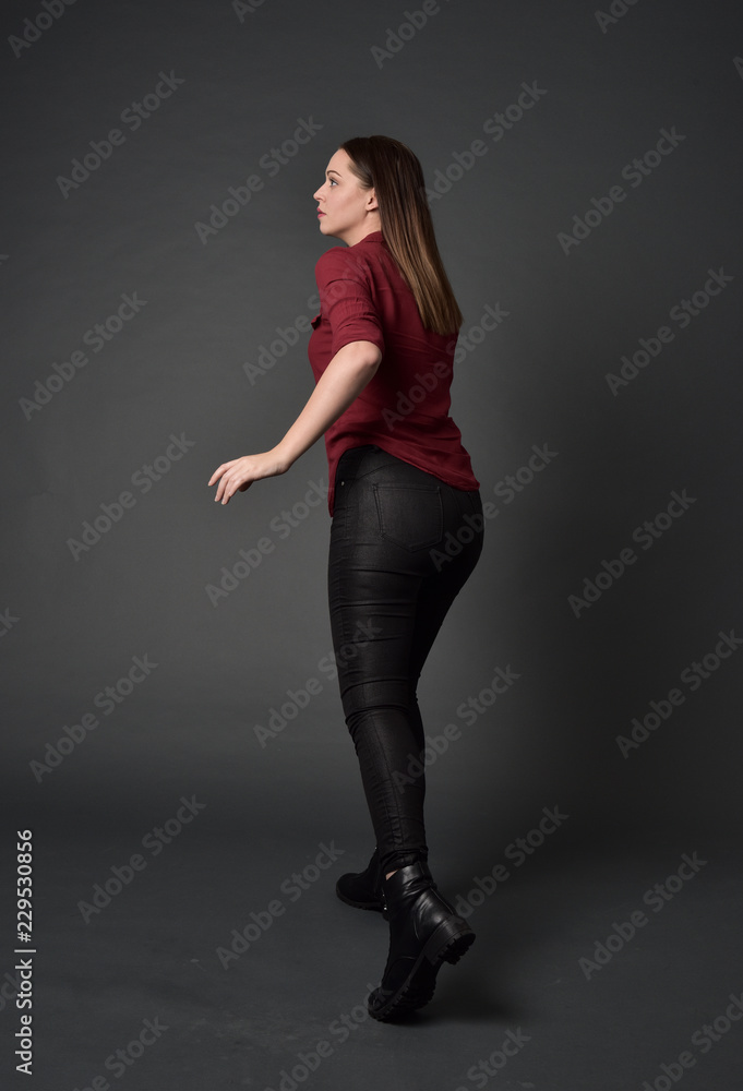 full length portrait of brunette girl wearing red shirt and leather pants. standing pose, facing away from the camera, on grey studio background. full length portrait of brunette girl wearing red shirt and leather pants. standing pose, facing away from the camera, on grey studio background.