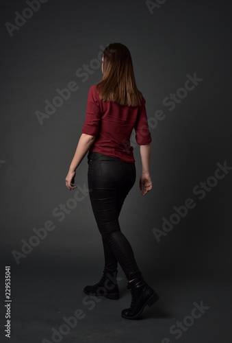 full length portrait of brunette girl wearing red shirt and leather pants. standing pose, facing away from the camera, on grey studio background.