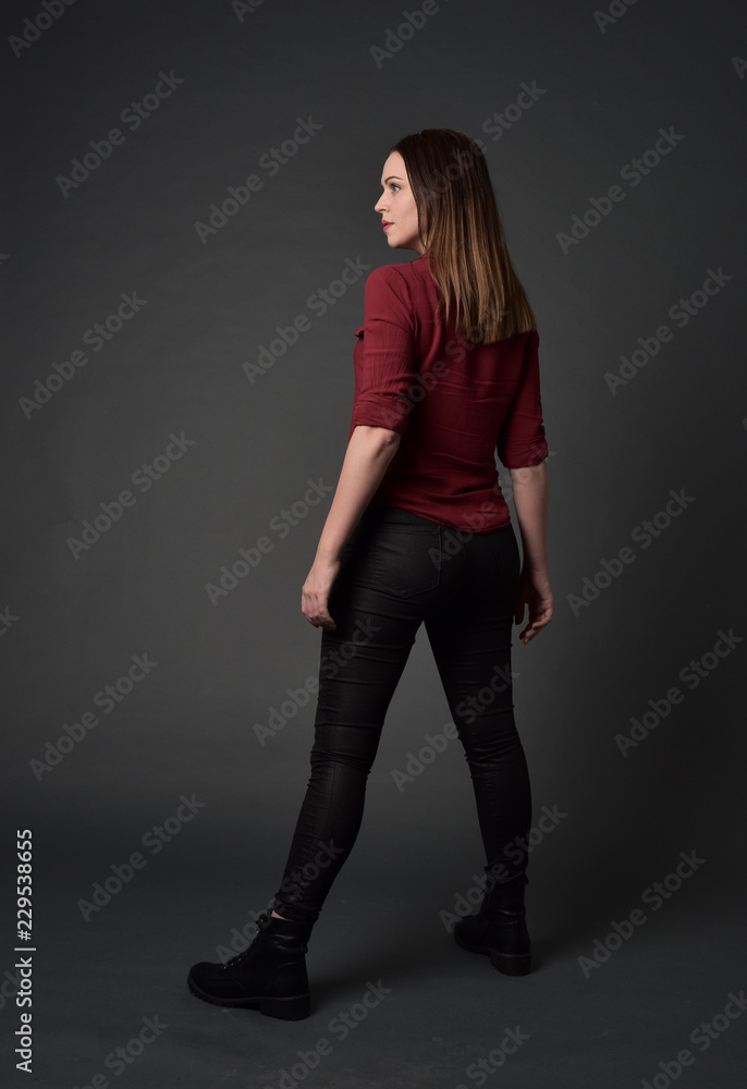 full length portrait of brunette girl wearing red shirt and leather pants. standing pose, facing away from the camera, on grey studio background. full length portrait of brunette girl wearing red shirt and leather pants. standing pose, facing away from the camera, on grey studio background.