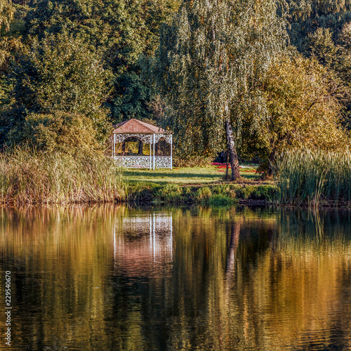 Carved wooden white gazebo with red roof by the lake reflected in water in the park, golden hours.