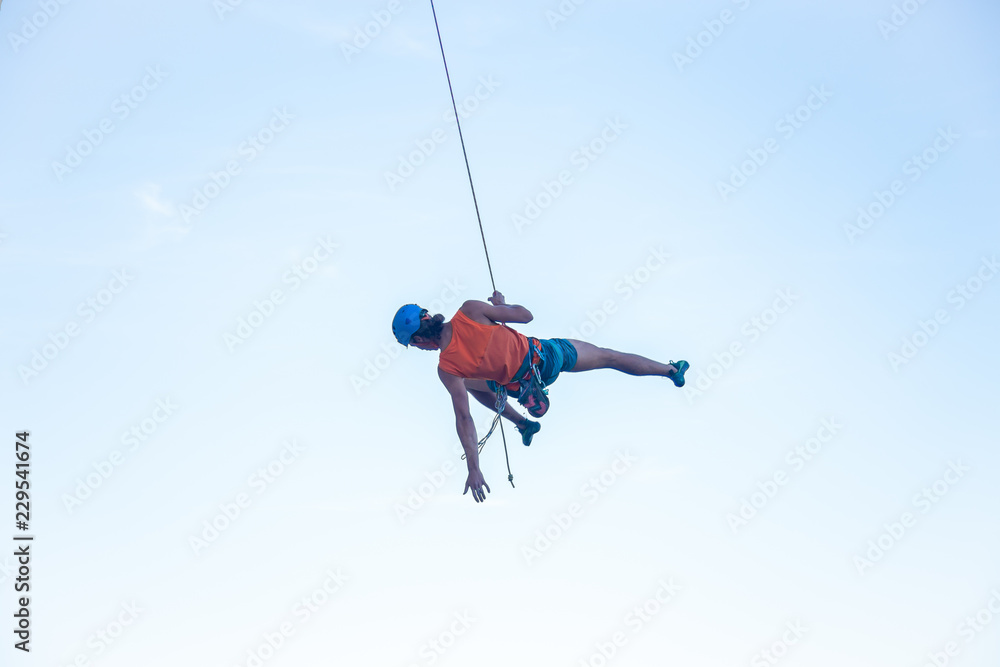 View of man in hardhat hanging on rope while doing rappel and showing pirouettes flying in air View of man in hardhat hanging on rope while doing rappel and showing pirouettes flying in air