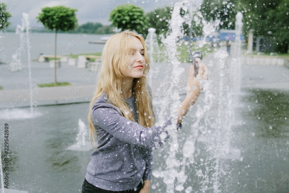 Fototapeta premium Portrait of beautiful student teenager girl holding up a smartphone taking selfies photos, networking using technology, outdoors park. Adolescent with books near water fountain, lifestyle exterior.