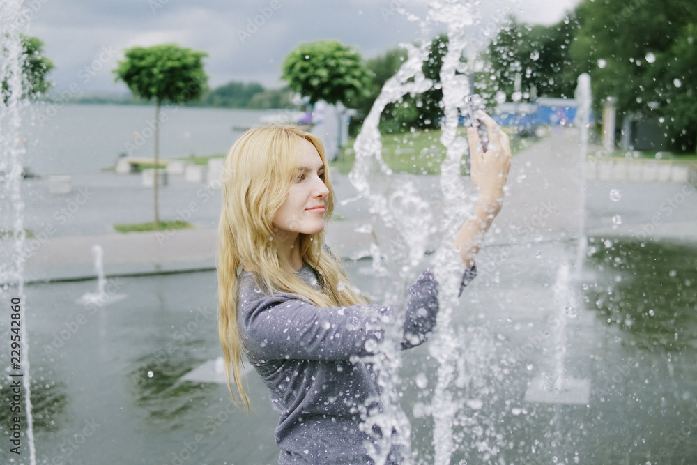 Obraz premium Portrait of beautiful student teenager girl holding up a smartphone taking selfies photos, networking using technology, outdoors park. Adolescent with books near water fountain, lifestyle exterior.