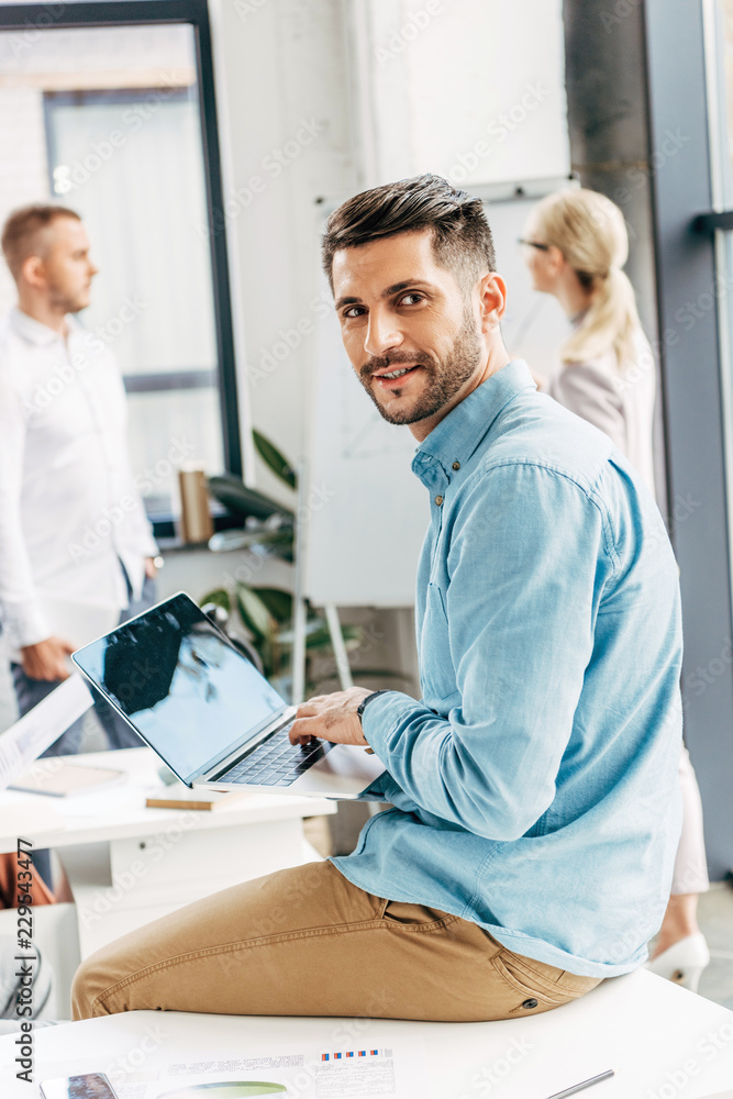 Fototapeta premium handsome young businessman using laptop and smiling at camera in office