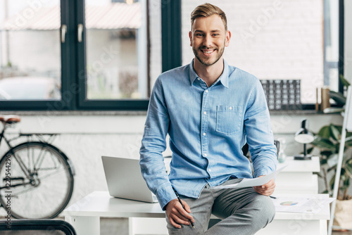 handsome young businessman holding papers and smiling at camera in office photo