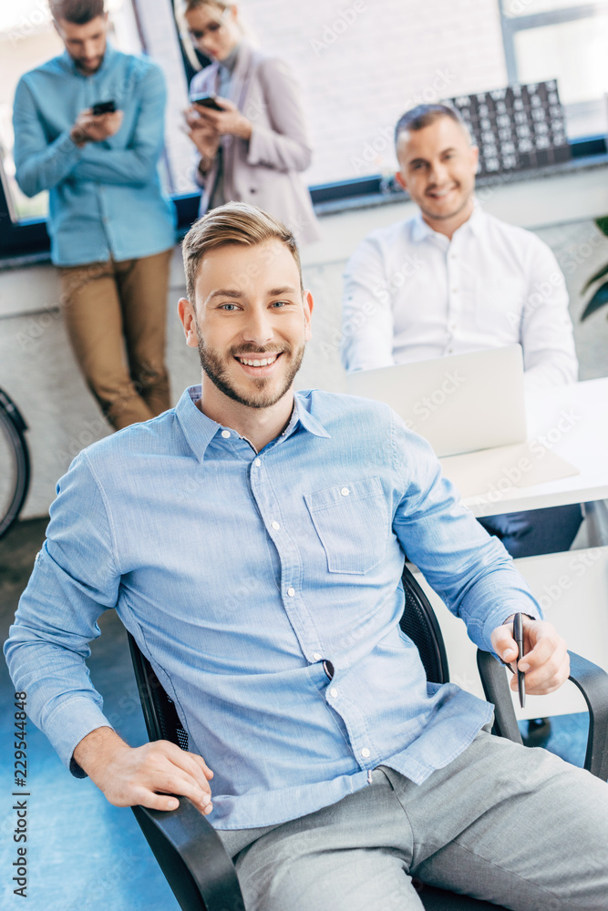 Fototapeta premium handsome young businessman smiling at camera while working with colleagues in office