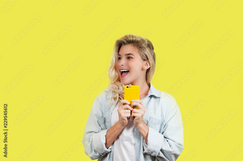 emotional very beautiful young girl looks into the phone and celebrates the victory standing on an isolated yellow background.