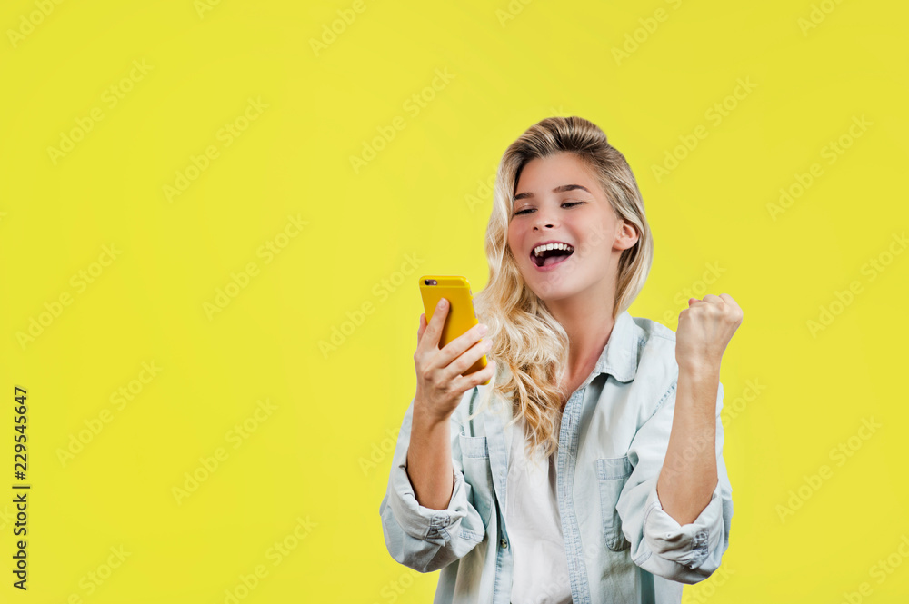 emotional very beautiful young girl looks into the phone and celebrates the victory standing on an isolated yellow background. emotional very beautiful young girl looks into the phone and celebrates the victory standing on an isolated yellow background.