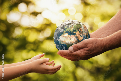 Fototapeta Naklejka Na Ścianę i Meble -  Close up of senior hands giving small planet earth to a child over defocused green background with copy space