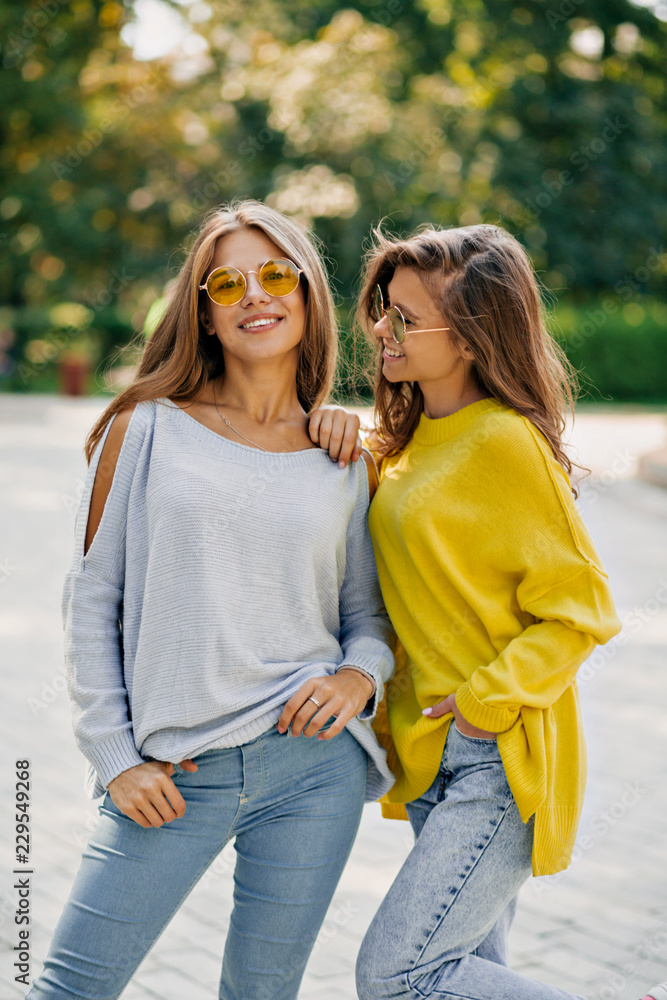 Fototapeta premium Two happy funny hipster girls in bright glasses and pullovers, laughing and sensing kiss, spend free time outside, bright summer outfits and accessories, listening music at headphones.