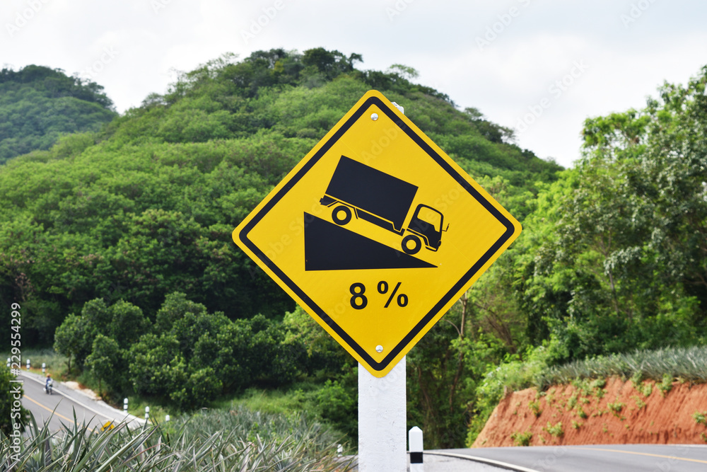 Traffic road sign warning down to hill steep climb (8%) , Paved road in the forest filled with trees and pineapple plantation at mountain in Thailand
