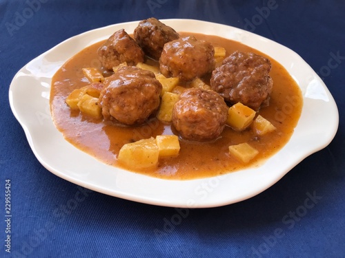 meatballs cooked in sauce of vegetables with a lining of potatoes. Typical Spanish food in white plate and blue background