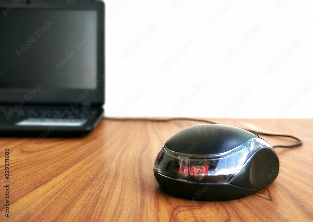 Focus on black laptop mouse with red light connecting on wooden table and blur laptop with white wall in background on technology equipment concept Focus on black laptop mouse with red light connecting on wooden table and blur laptop with white wall in background on technology equipment concept