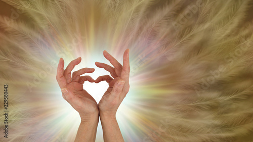 Sending out unconditional love healing vibes - hands making a love heart symbol with a white burst of light behind and a gold brown feather effect background with copy space
 photo