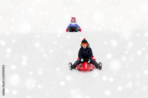 childhood, sledging and season concept - happy little kids sliding on sleds down snow hill in winter