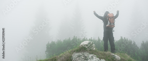 Hiking man with backpack open arms on foggy mountain peak