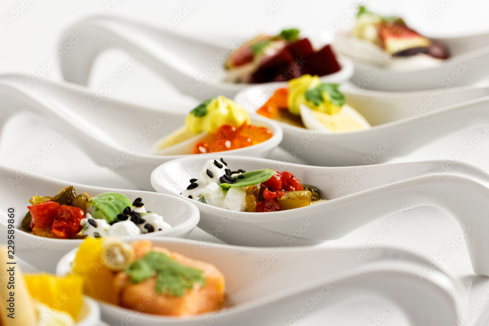 A row of mixed canapes in white ceramic spoons on white background.