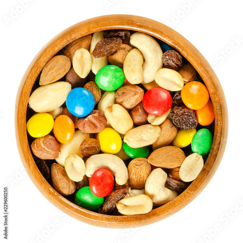 Trail mix in wooden bowl. Snack mix. Almonds, cashews, peanuts, hazelnuts, raisins and colorful chocolate candies. Food to be taken along hikes. Macro food photo closeup from above on white background