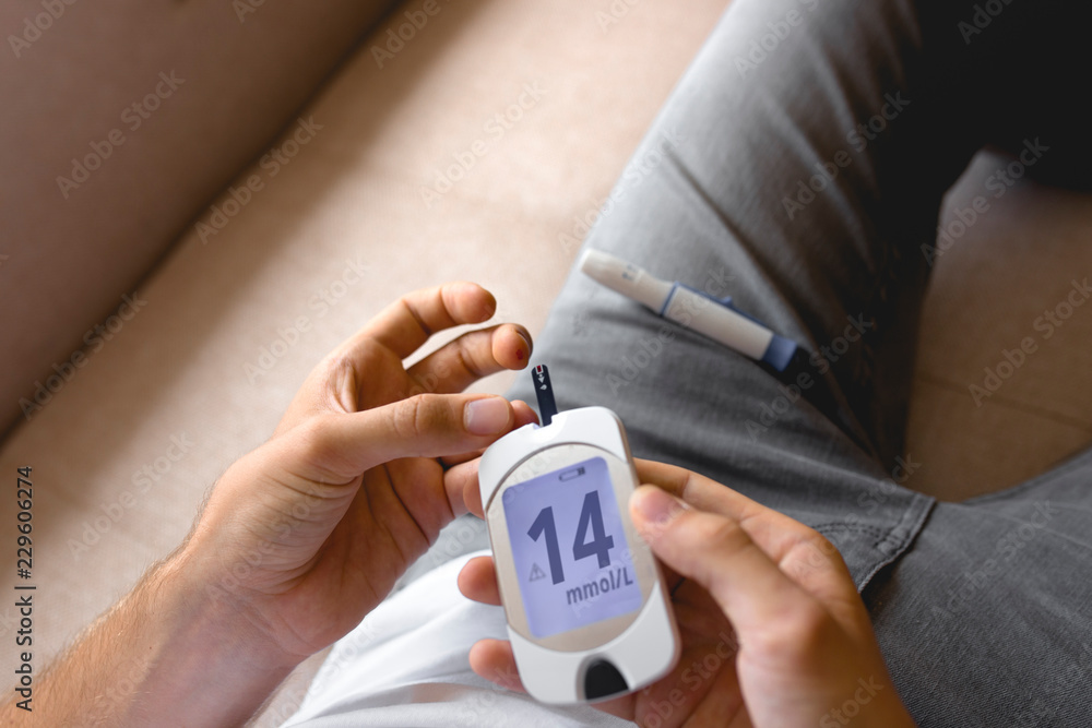 Fototapeta premium Handsome stylish young teen caucasian white brunette guy man european taking a blood sample with a glucometer to measure sugar level,lancet on finger diabetes, glycemia meter top first view shot