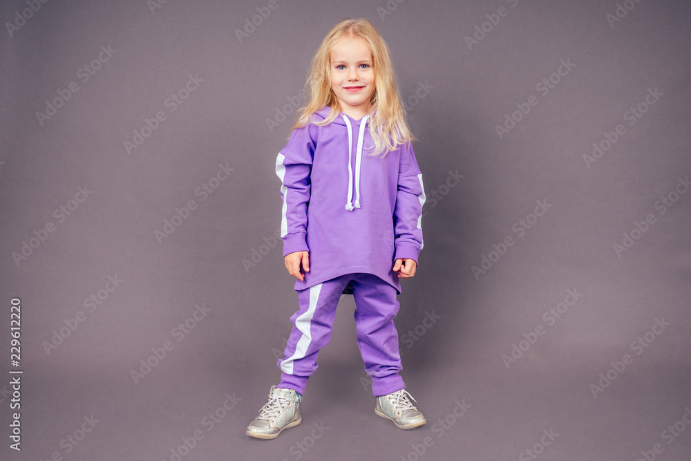 blonde blue-eyed little model girl in a purple sports suit posing in the studio on a black background.active childhood full length portrait violet sporty costume blonde blue-eyed little model girl in a purple sports suit posing in the studio on a black background.active childhood full length portrait violet sporty costume