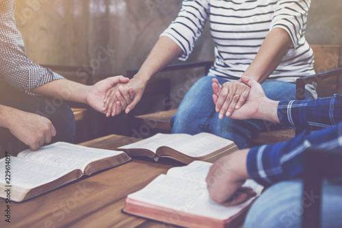 christian small group holding hands and praying together around wooden table with blurred open bible page in home room, devotional or prayer meeting concept