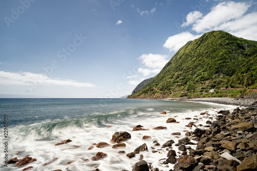 Lonely beach with big boulders, a wave rolls up, water movement in long exposure, mountain peak with lush vegetation in the background, depth effect - Location: Portugal, Azores, island of Sao Jorge