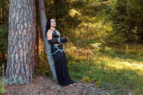 A young witch in black clothes is chained to a tree. Halloween. Horizontal image.