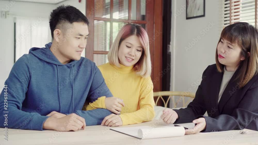 Happy young Asian couple and realtor agent. Cheerful young man signing some documents while sitting at desk together with his wife. Buying new house real estate. Signing good condition contract. Happy young Asian couple and realtor agent. Cheerful young man signing some documents while sitting at desk together with his wife. Buying new house real estate. Signing good condition contract.