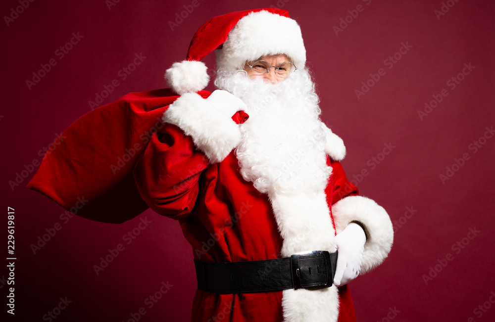 Portrait of grandfather in Santa Clause costume with gift sack on back posing on red background, Christmas and New year concept