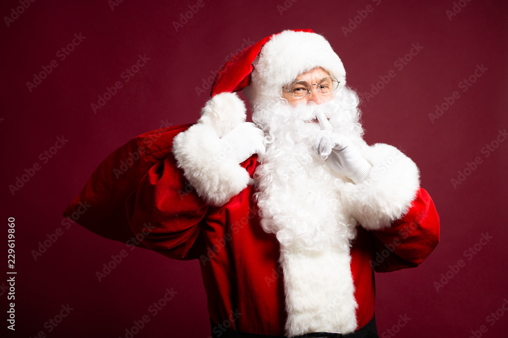 Portrait of man in Santa Clause costume looking at camera and showing silence sign on red background, Christmas and New year concept