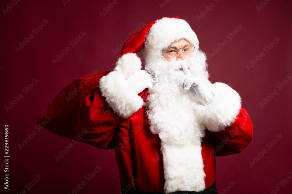 Portrait of man in Santa Clause costume looking at camera and showing silence sign on red background, Christmas and New year concept Portrait of man in Santa Clause costume looking at camera and showing silence sign on red background, Christmas and New year concept