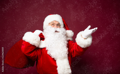 Portrait of grandfather in Santa Clause costume with gift sack on back posing on red background, Christmas and New year concept