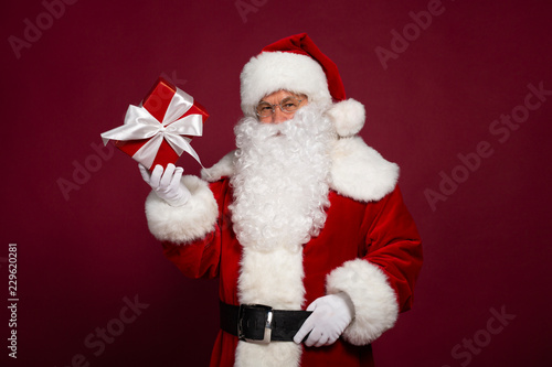 Portrait of man in Santa Clause costume holding paper gift box and looking at camera on red background, Christmas and New year concept
